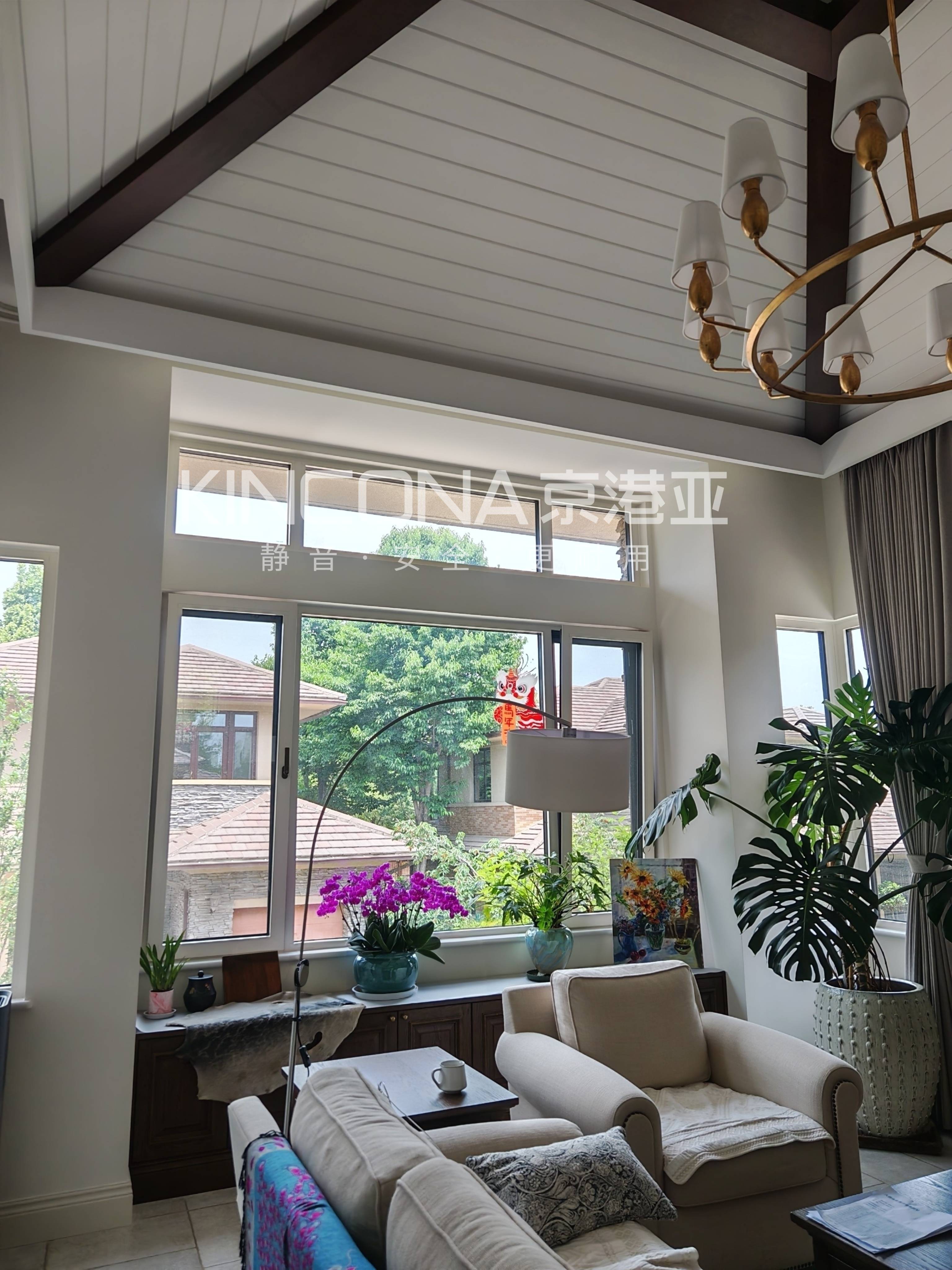 这是一张展示京港亚门窗实景案例的图片。画面为一个拥有高挑空斜坡顶的明亮客厅,大面积白色落地窗带来极佳采光,窗外可见绿树与红瓦建筑。室内陈设米色布艺沙发、深色木柜