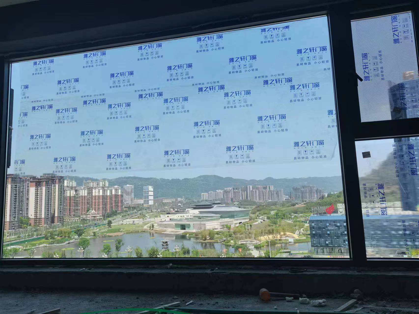 室内装修施工现场，黑色边框的雅之轩品牌大窗户已安装到位。玻璃表面贴满印有“雅之轩门窗”及“易碎物品 小心轻放”字样的蓝色保护膜，有效防止施工刮花。窗外视野极佳，